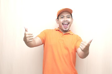 Naklejka premium Southeast Asian man in orange uniform and cap showing two thumbs up on beige background, expressing approval, success, and service satisfaction.