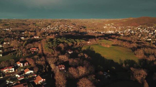 Aerial view of Ascain's landscape, where houses nestle amidst trees, bathed in warm sunlight contrasting with the cloudy sky, Ascain, Nouvelle-Aquitaine, France.