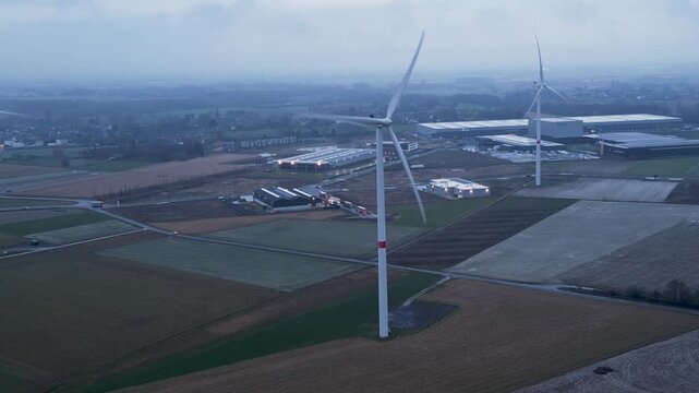 Aerial view of wind turbines standing tall in a landscape of fields and industrial warehouses, with a muted color palette, Tournai, Wallonia, Belgium.