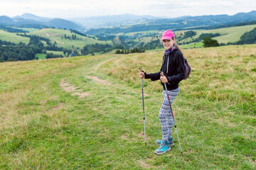 Young Female Hiker with Pink Cap, Patterned Leggings, Trekking Poles and Backpack on Grassy Hill overlooking Green Mountain Valley Panorama