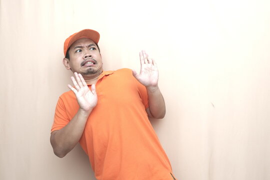 Southeast Asian man in orange cap and polo leans back with raised hands, showing fear, rejection, and avoidance on beige background.