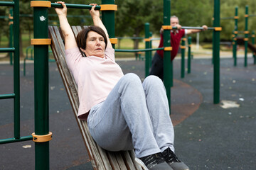 Fototapeta premium Mature woman goes in for sports in city park on sports ground - doing exercises to pull his legs to chest