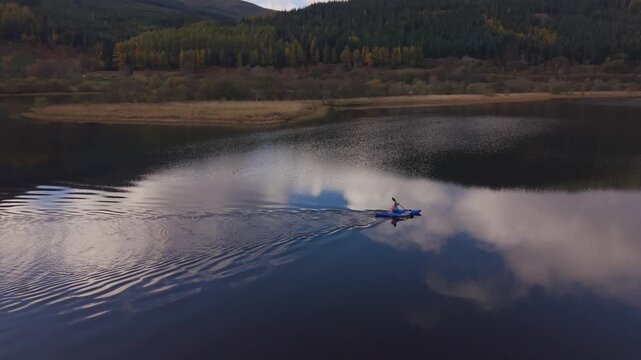 Aerial view of a lone kayaker cutting through the dark, reflective waters of a serene lake surrounded by colorful trees, Callander, Scotland, United Kingdom.