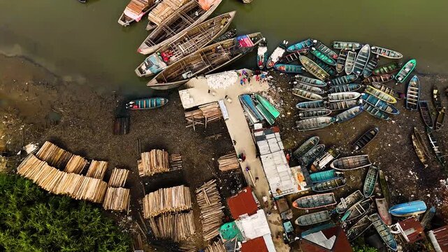 Aerial view of colorful boats lining the muddy riverbank next to a lumber yard, creating a vibrant contrast of industry and nature, Port Harcourt, Rivers State, Nigeria.