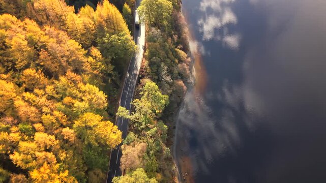 Aerial view of a road cutting through autumn foliage and reflecting the sky in a loch, offering a scenic view, Callander, Scotland, United Kingdom.