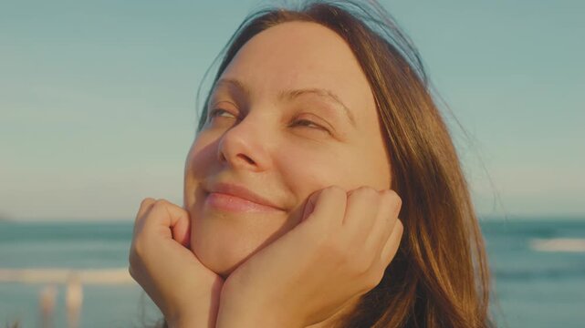 Woman smiling with hands cradling her chin, looking up towards the light, finding joy and contemplation on a serene beach with ocean waves in the background during golden hour