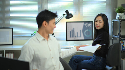 Colleagues exchange documents in a modern home office, discussing work progress with a computer and charts