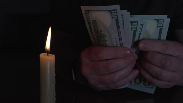 Man counting dozens of dollars bills by candlelight in a dark room, hands and cash half-illuminated by a flickering flame suggesting secrecy, risk, and illicit finance