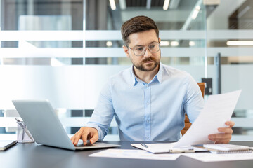 Obraz premium Businessman in glasses and blue shirt performing data analysis, reviewing important paperwork, and typing on a computer, demonstrating efficiency and focus in a modern corporate workplace