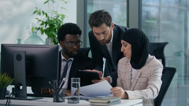 Diverse business team arguing in office angry discussion documents computer work conflict Muslim woman in hijab African Caucasian male colleagues stressed teamwork problem multiracial three co-workers