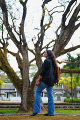 Woman with wavy hair and backpack standing outdoors on a pathway, looking up at the large, ancient tree branches, contemplating nature and exploration
