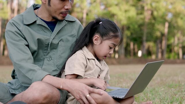 Asian adult father and young daughter sitting closely using laptop together in forest park grass area focusing on learning during digital education moment while enjoying peaceful family vacation outdo