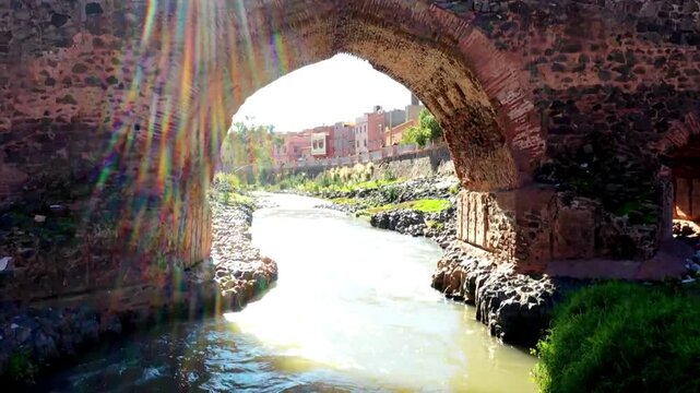 HD shot of Oum Er-Rbia river flowing through Khenifra city viewed through a stone archway. Features flowing water, rocky banks, and urban architecture in the Middle Atlas.