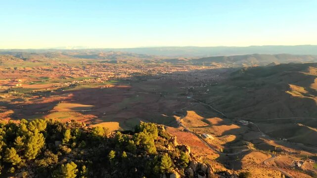 Wide aerial drone shot of rolling hills and red earth landscape in the Middle Atlas. Features rugged terrain, sparse vegetation, and scenic mountain geography in Morocco.