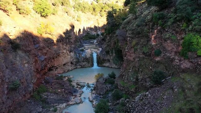Aerial drone shot of the Oum Er-Rbia river source waterfall cascading into a turquoise pool. Features rugged rocky cliffs and natural springs in the Middle Atlas mountains.