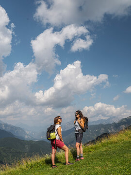 Two women with backpacks stand on a grassy hillside, gazing at a mountain range under a blue sky with puffy clouds. Casera Coot,Resia Valley,Julian Alps,Italy