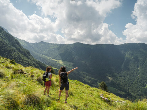 Two hikers with backpacks stand on a grassy hillside, one pointing towards a valley and mountains. Casera Canin,Resia Valley,Julian Alps,Italy