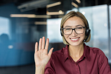 Young woman wearing a headset and glasses, waving brightly while smiling at the camera, providing...