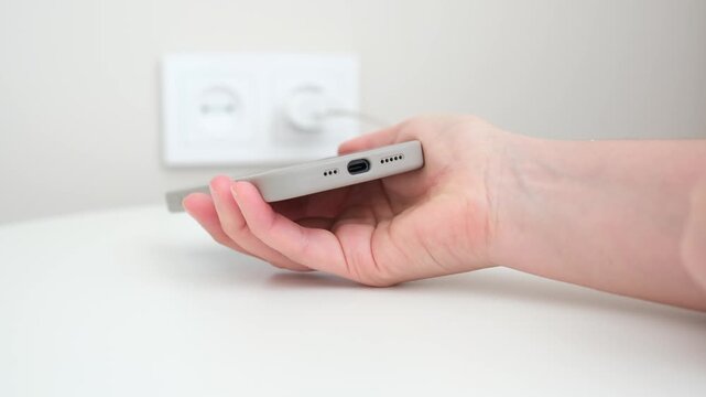White Hand Plugging Cable Into Battery, CloseUp On Fingers Inserting USB Connector On White Tabletop Near Wall Outlet, Neutral Background, Indoor Home Scene, Minimalist