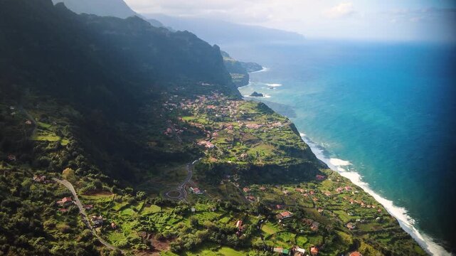 Establishing shot of Arco de Sao Jorge village from Beira da Quinta viewpoint in Madeira, aerial reveal of coastal cliffs and traditional red roofs under bright sunlight, mountain landscape.