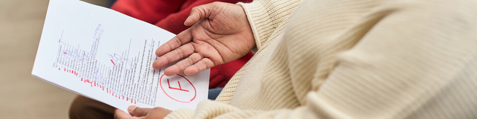 Header of Middle aged black woman holding failed test paper with large red F grade, gesturing with hand while discussing result, partial view of child in background © DragonImages