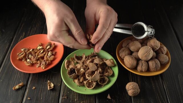 Hands of a person cracking open walnuts and separating the shells from the nuts on a wooden table with colorful plates in a kitchen setting