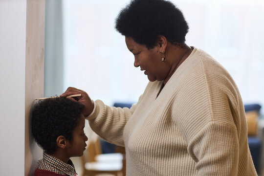 Black middle aged woman measuring height of Black child boy standing against wall, using hand and pencil for marking, both focused on process, indoor family setting