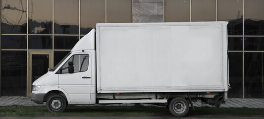 Side view of delivery truck with large blank white panel on its cargo area parked on side of road. Truck is slightly worn and urban background includes modern office glasses  buildings © Maxim Chuev