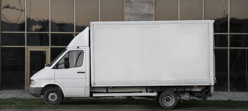 Side view of delivery truck with large blank white panel on its cargo area parked on side of road. Truck is slightly worn and urban background includes modern office glasses  buildings