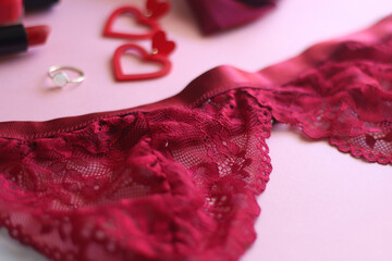 Red lace bralette, hairbow, heart shaped earrings, golden ring, red lipstick and nail polish on pink background. Coquettish accessories for Valentine's day. Selective focus.