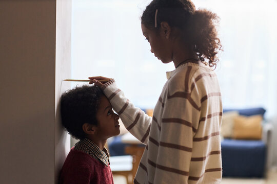 Black girl teenager measuring height of Black boy child against wall using pencil, both standing indoors, girl focusing on marking while boy looking up at her with anticipation
