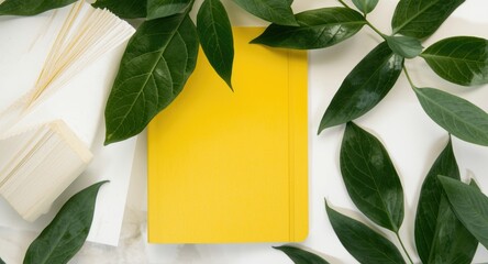 Flat lay composition of a desk featuring a yellow notebook and vibrant green leaves on a white background