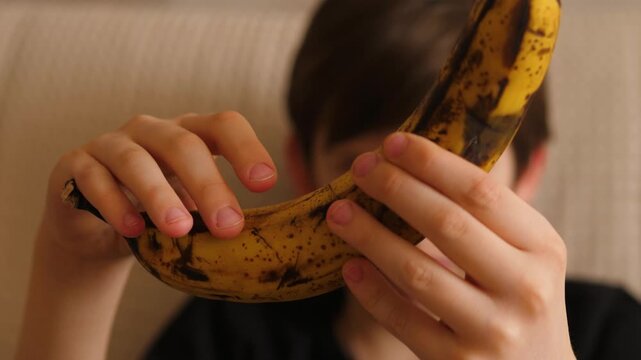 A teenager examines an old overripe banana with brown spots with distrust and disgust. 