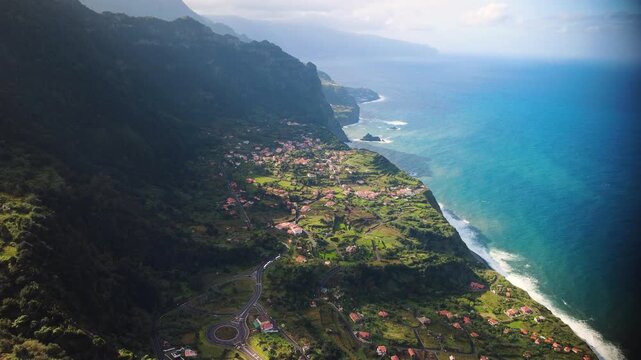 Establishing shot of Arco de Sao Jorge village from Beira da Quinta viewpoint in Madeira, aerial reveal of coastal cliffs and traditional red roofs under bright sunlight, mountain landscape.