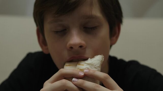A young boy is eating a delicious shawarma wrap with great pleasure. This close-up shot captures the child's happy emotions as he enjoys his tasty snack with eyes closed