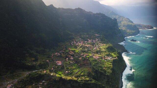 Aerial tracking shot of Arco de Sao Jorge village in Madeira, drone flying sideways revealing massive mountains and Atlantic ocean with powerful crashing waves, cinematic coastal establishing shot.