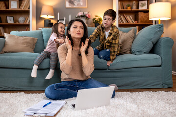 Exhausted mother trying to focus on laptop while kids demand attention. Remote work challenges and parenting stress. Annoyed woman working from home with energetic children in the background.