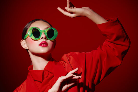 Fashion model wearing oversized green sunglasses and bold red lipstick poses against red background in a stylized beauty creative concept involving vivid colors and dramatic lighting.