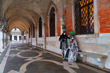 Venetian carnival couple wearing matching historical costumes and elegant masks, standing together against the historical architecture of Venice