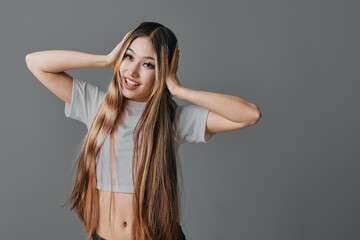 Young woman with long hair in casual white shirt smiling and holding her head with both hands against gray background. Portrait of happy female showing natural emotions and style.