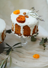 Handmade Easter cakes with glaze and dried citrus slices decorated with green leaves on bright textile background. Minimal light food photography style.