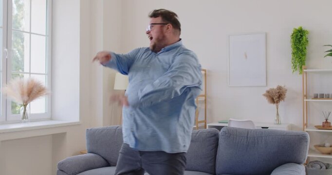 Expressive fat overweight man in glasses dancing and singing with upbeat energy near a sofa in living room, enjoying music and the moment at home, carefree. Obese male person having fun indoors.