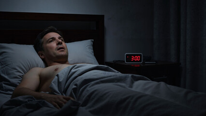 Obraz premium Caucasian man experiencing insomnia late at night, unable to sleep with an alarm clock showing 3:00 am next to his bed. Sleep disorder concept.