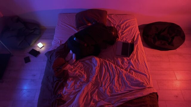 Lonely young man curled up on bed at night with phone and laptop in colorful neon light, emotional exhaustion, overthinking and quiet sadness in modern digital life
