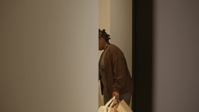 Tired African American woman entering flat, carrying paper bags. Her husband smiling to her and helping with purchase