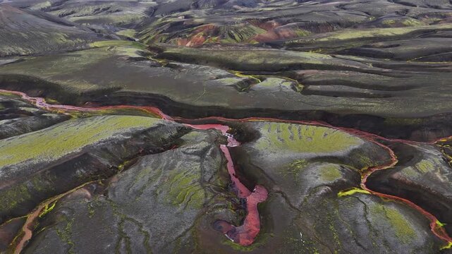 Aerial view of a striking red river cutting through a dramatic landscape of dark, textured earth and vibrant green fields, Selfoss, Iceland.