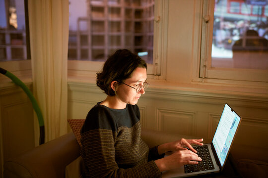 Woman with glasses typing on a laptop in a cozy room with city view windows. Belgium
