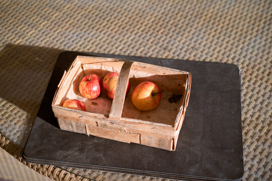 Wooden crate with red apples on a woven wicker table surface under sunlight. Germany