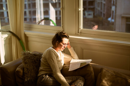 Woman reading a document on a sofa bathed in warm sunlight from a window. Belgium