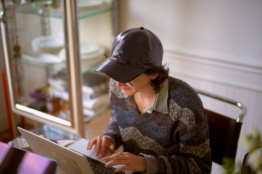 Young adult wearing a cap uses a laptop at a glass table in a warmly lit room. Belgium